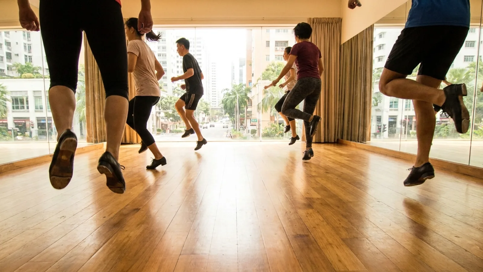 A row of dancers in tap shoes mid-step on a wooden studio floor during a tap dance class in a bright Singapore dance studio