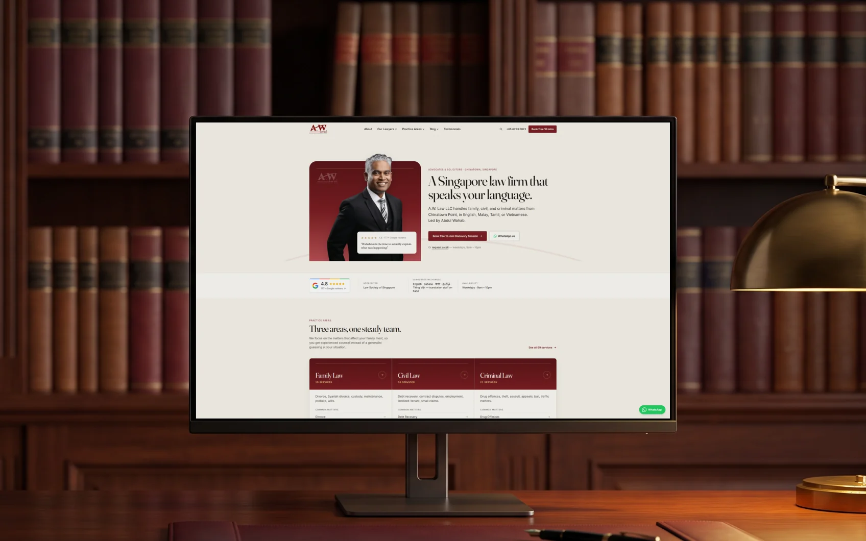 A.W. Law LLC homepage, "A Singapore law firm that speaks your language." beside Abdul Wahab's portrait, on a 27-inch monitor on a mahogany desk in a wood-panelled office with a brass lamp, in warm evening light
