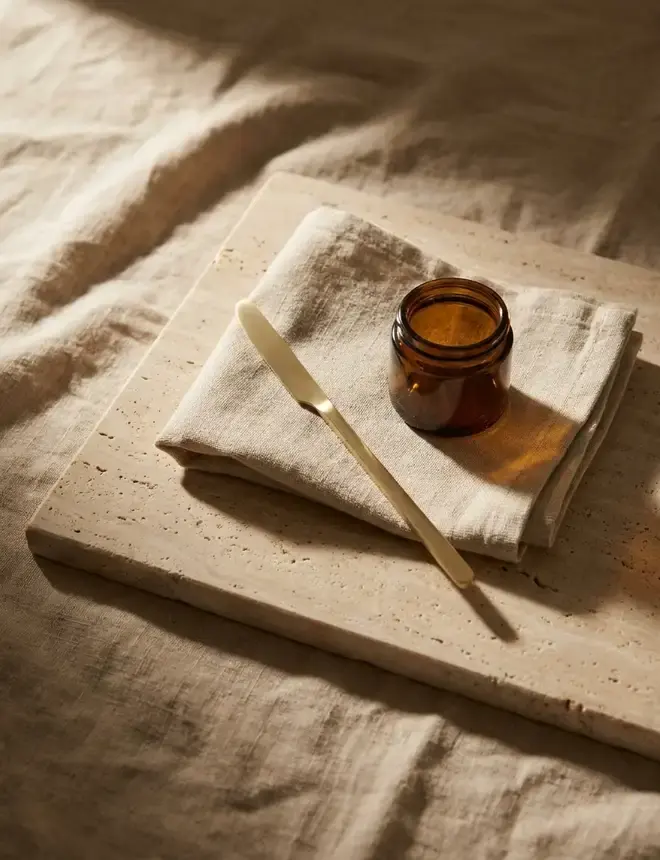 Still life of travertine, folded oat cloth, a brushed brass tool and an amber glass vessel on raw linen