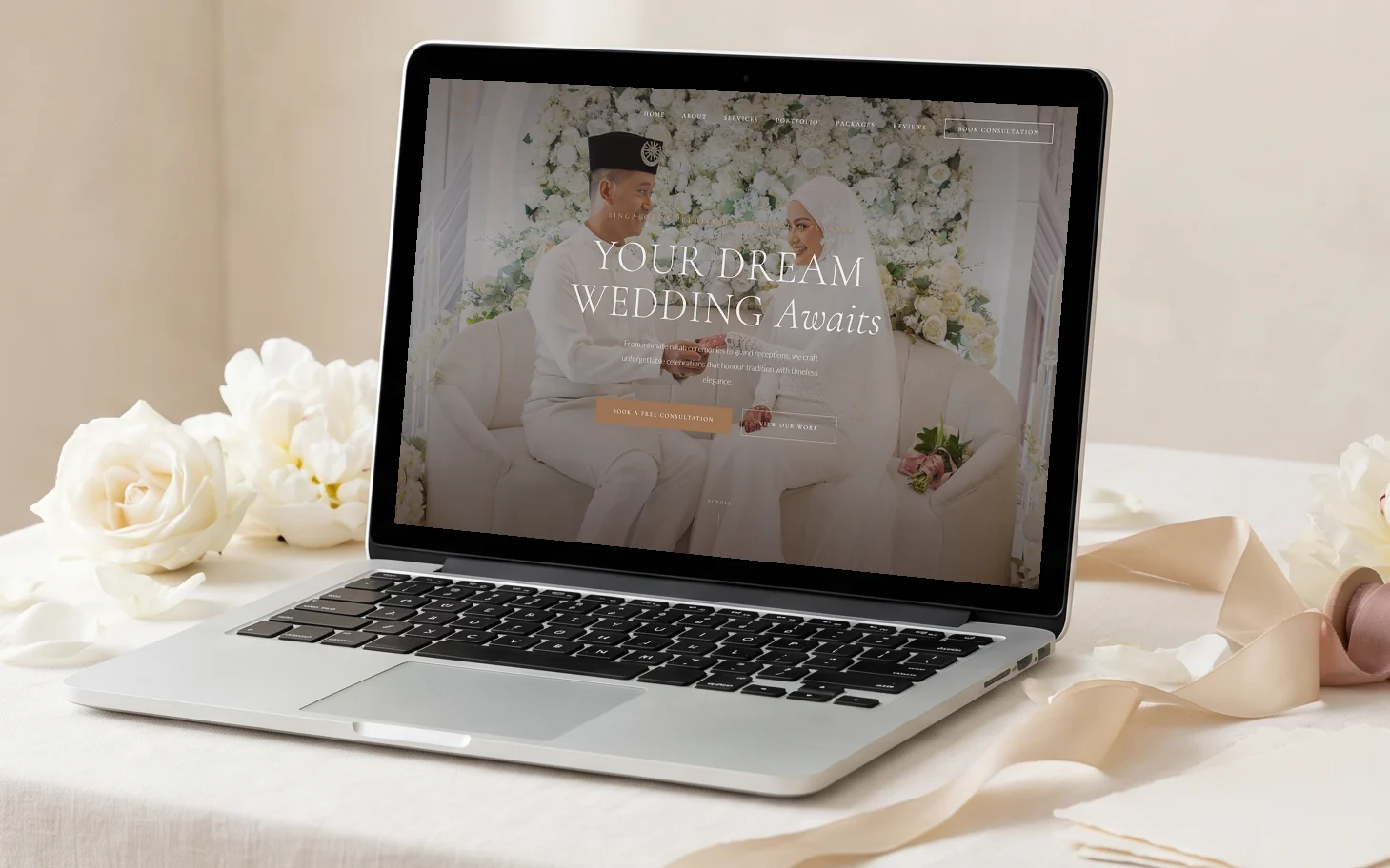 GFF Weddings homepage concept, "Your Dream Wedding Awaits" over a Malay bride and groom at their floral pelamin, on a silver laptop on an ivory linen wedding-planning table with white roses and peonies, silk ribbon and blank invitation cards