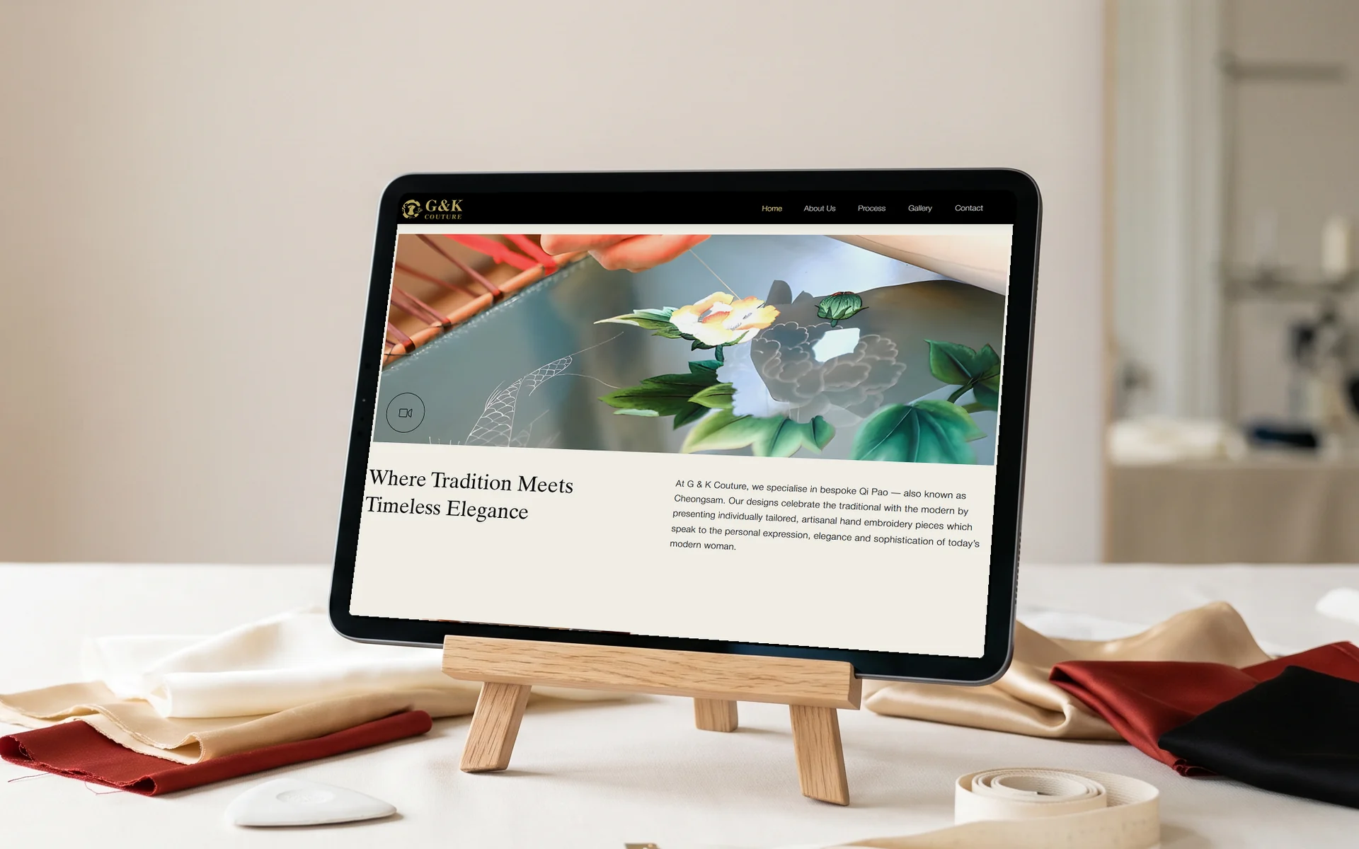 G & K Couture homepage, "Where Tradition Meets Timeless Elegance" beneath its hand-embroidery hero, on a tablet on a small oak easel at an atelier worktable with silk swatches, tailor's chalk and a measuring tape in soft window light