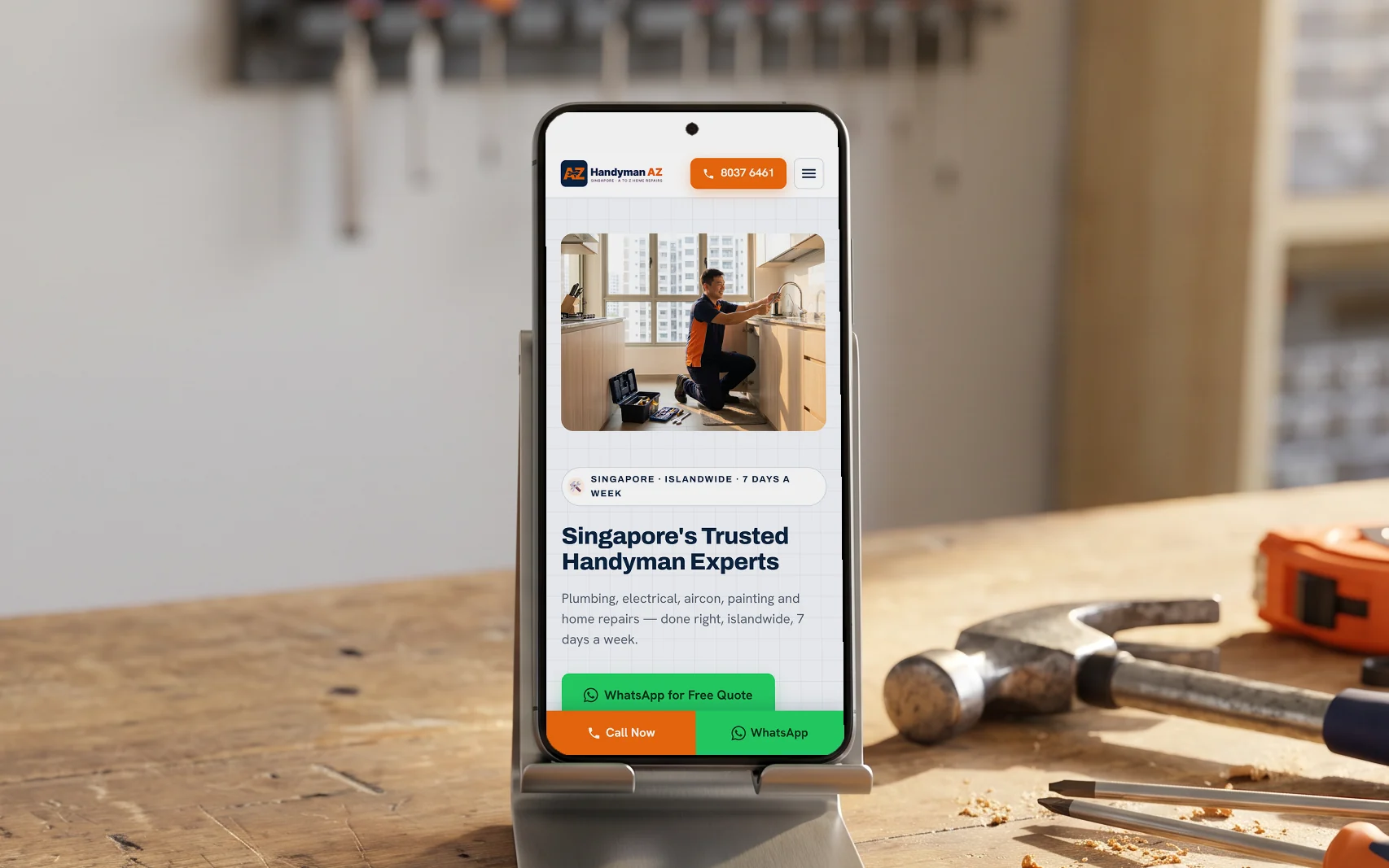 Handyman AZ mobile site, "Singapore's Trusted Handyman Experts" with its Call Now and WhatsApp bar, on a phone in a steel stand on a wooden workbench beside a hammer, tape measure and screwdrivers, in warm garage daylight