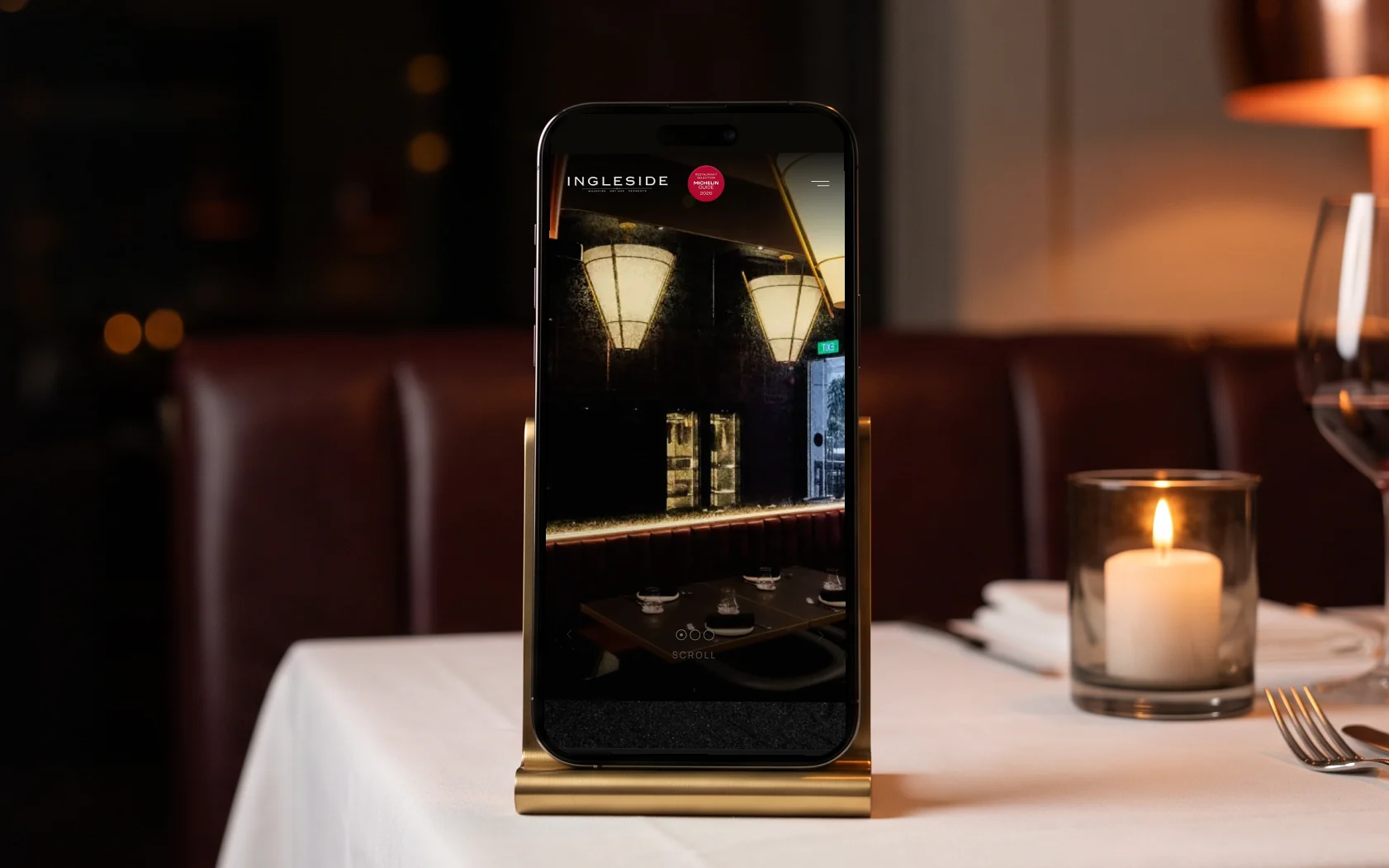 Ingleside mobile site, the Ingleside wordmark and its Michelin Guide 2026 badge over the dining room's cone pendants and black marble, on a phone in a brass stand on a candlelit table with white linen, a wine glass and cutlery