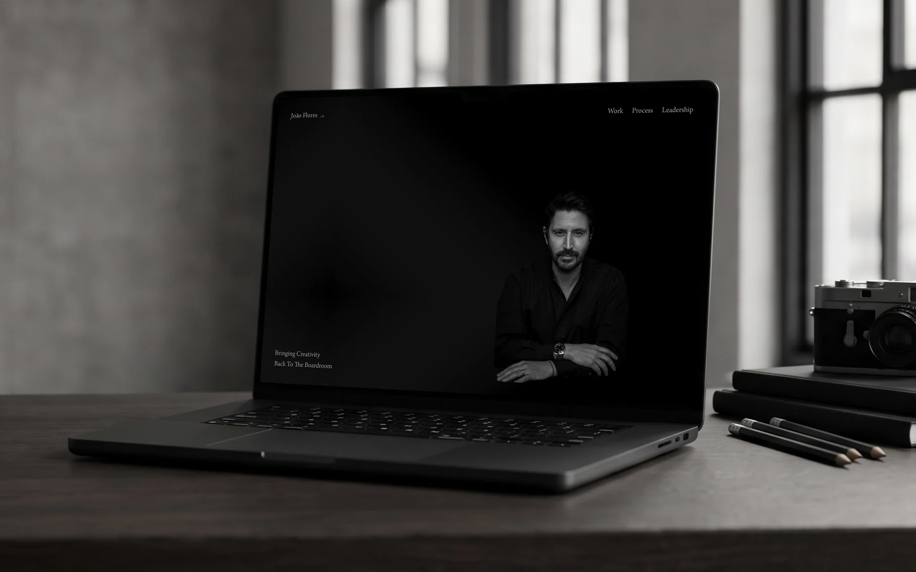 João Flores homepage, "Bringing Creativity Back To The Boardroom" beside a black-and-white portrait on black, on a laptop on a dark oak desk in a loft studio with sketchbooks and a film camera, in window daylight