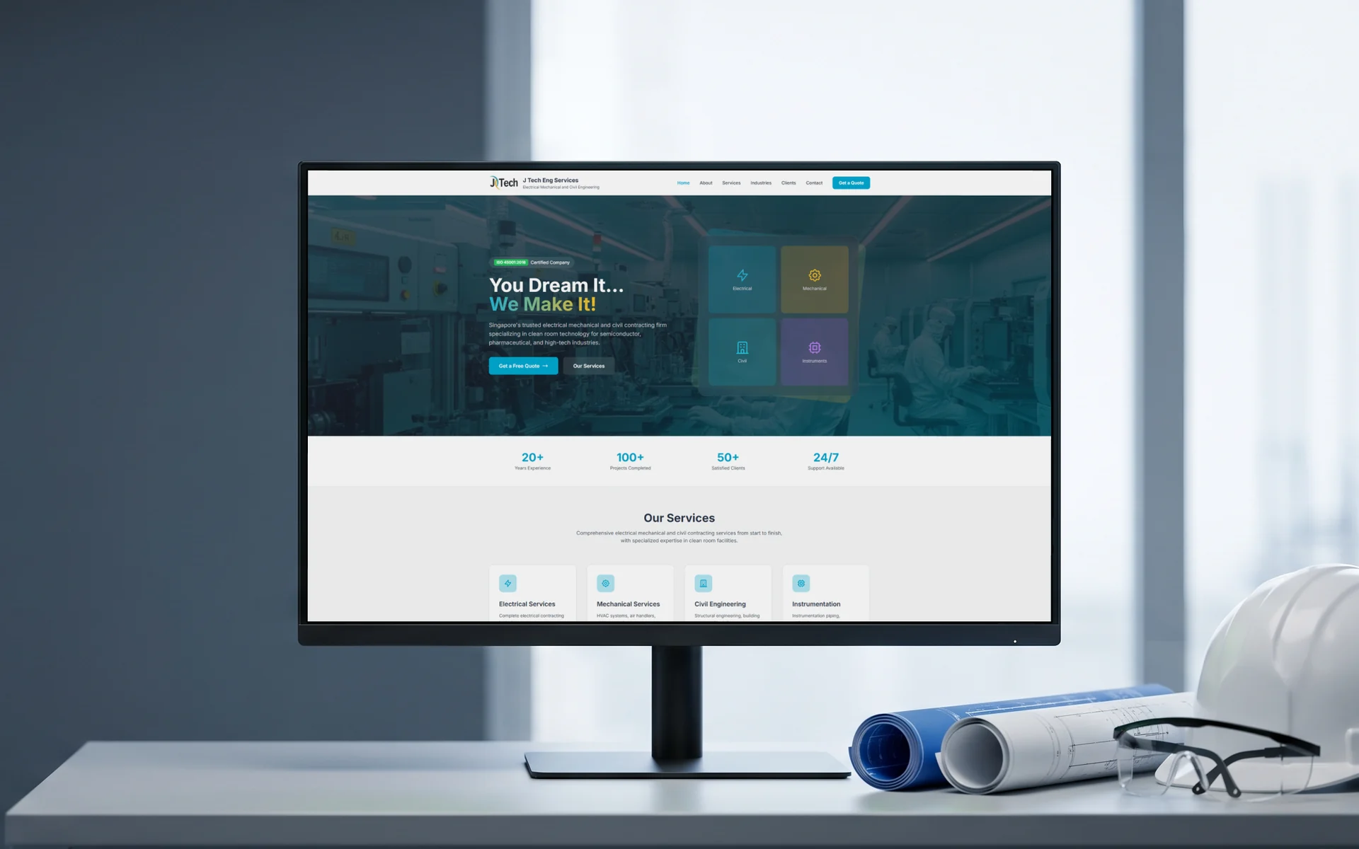 J Tech Eng Services homepage, "You Dream It... We Make It!" over a clean room photo, on a 27-inch monitor on a site office desk beside rolled blueprints, a white hard hat and safety glasses