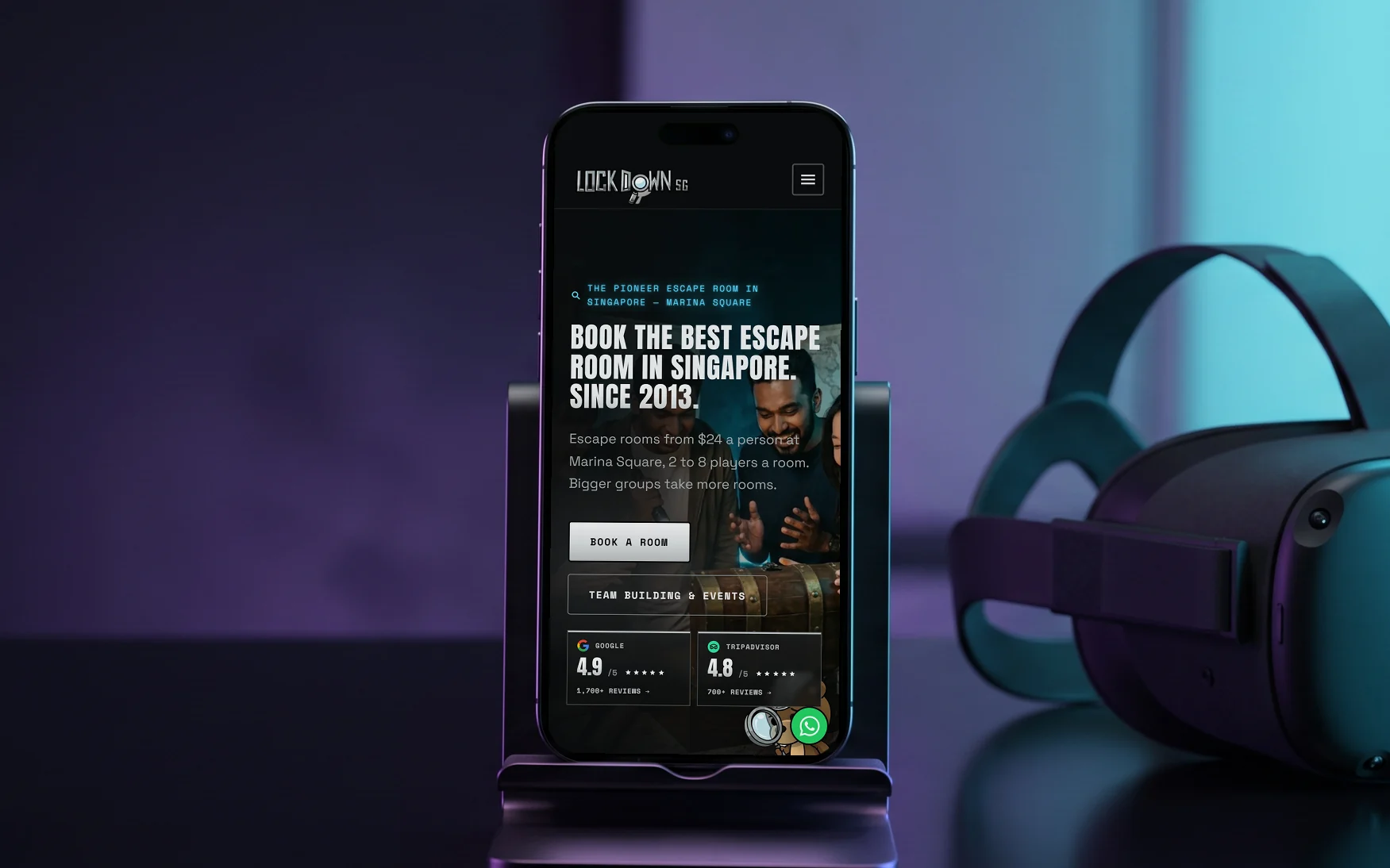 Lockdown.SG mobile site, "Book the best escape room in Singapore. Since 2013.", on a phone in a slim stand on a dark glossy counter beside a VR headset, in violet and cyan light