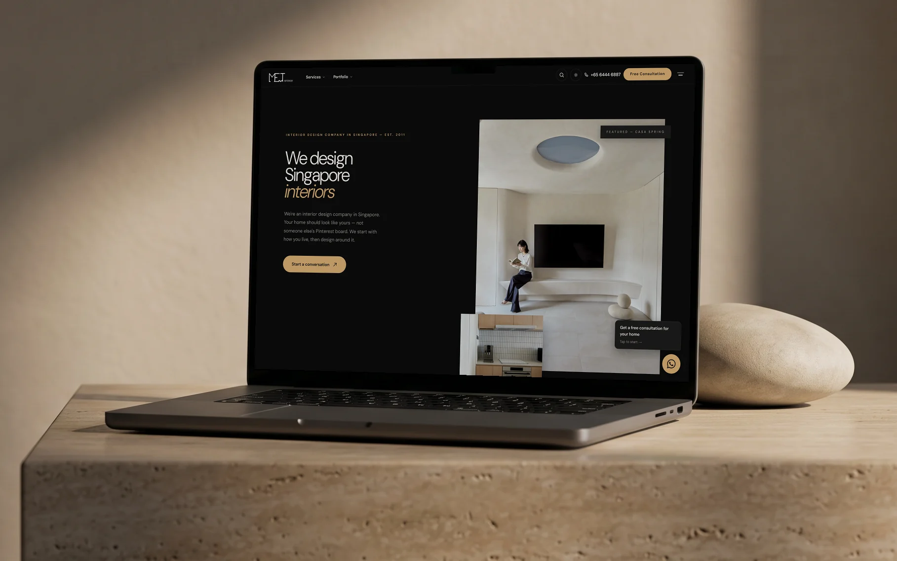 MET Interior homepage, "We design Singapore interiors" in dark mode, on a laptop resting on a travertine plinth beside a stone pebble