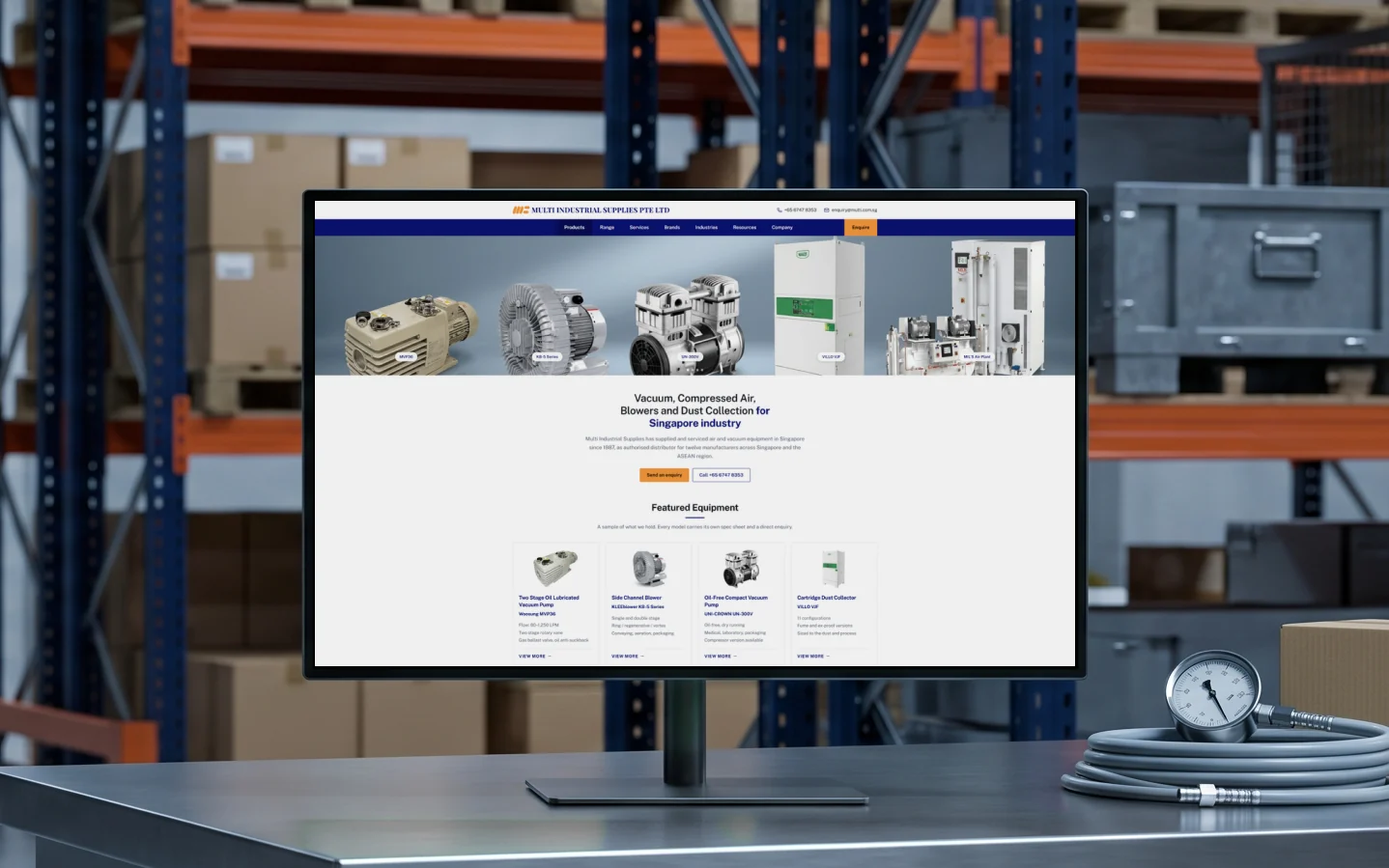 Multi Industrial Supplies homepage concept, "Vacuum, Compressed Air, Blowers and Dust Collection for Singapore industry" under a banner of five equipment models, on a monitor on a steel trade counter in a warehouse, navy and orange pallet racking soft behind
