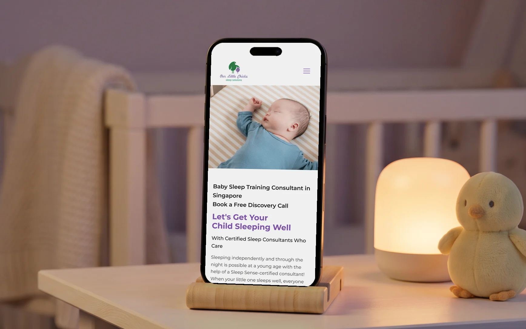 Our Little Chicks mobile site, "Let's Get Your Child Sleeping Well" under a photo of a sleeping baby, on a phone in a wooden stand on a nursery bedside table beside a glowing night light and a plush chick, in warm evening light