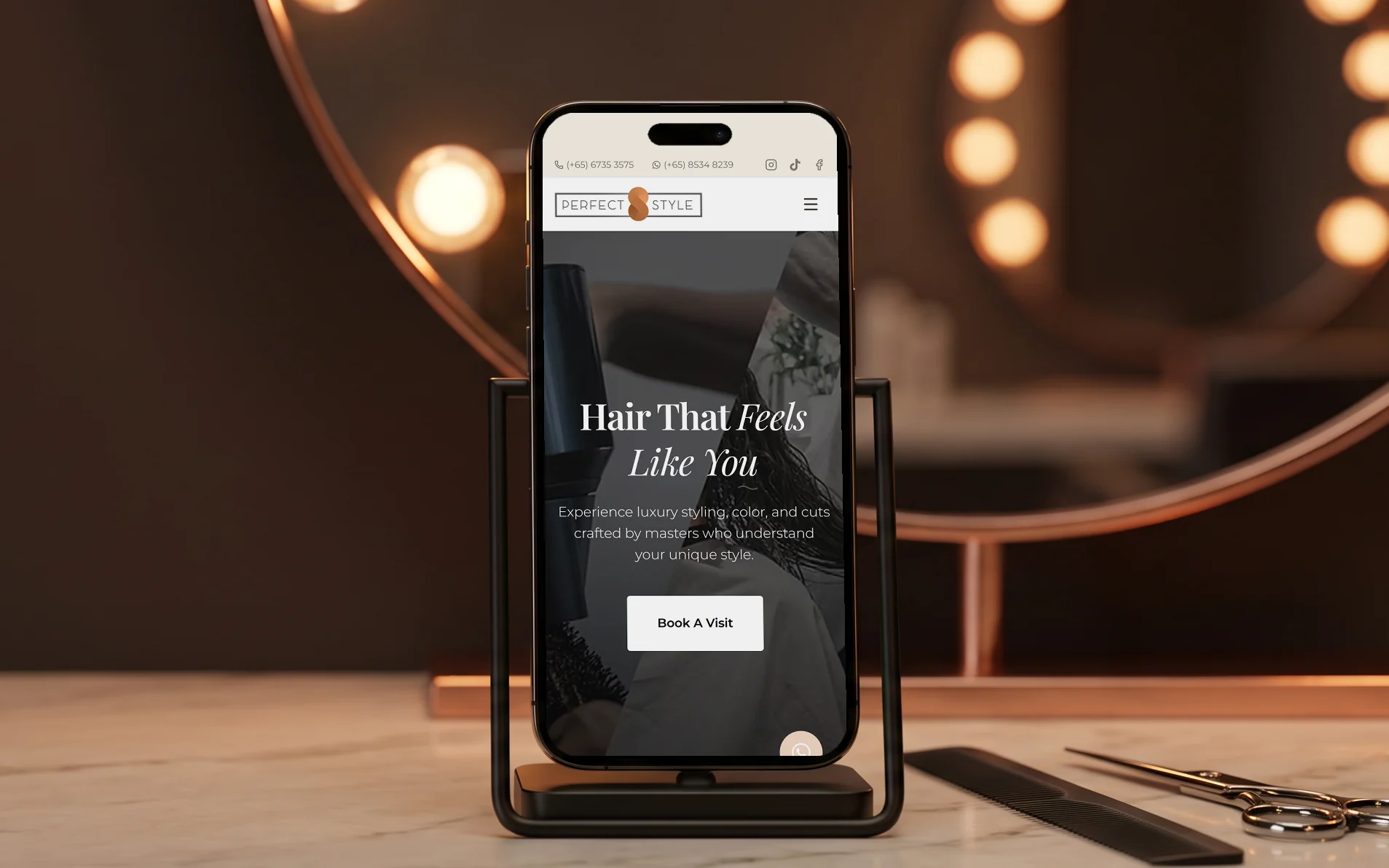 Perfect Style Salon mobile site, "Hair That Feels Like You" with its Book A Visit button, on a phone in a slim black stand at a salon styling station, on a marble counter beside a comb and shears, a round mirror ringed with vanity bulbs glowing behind