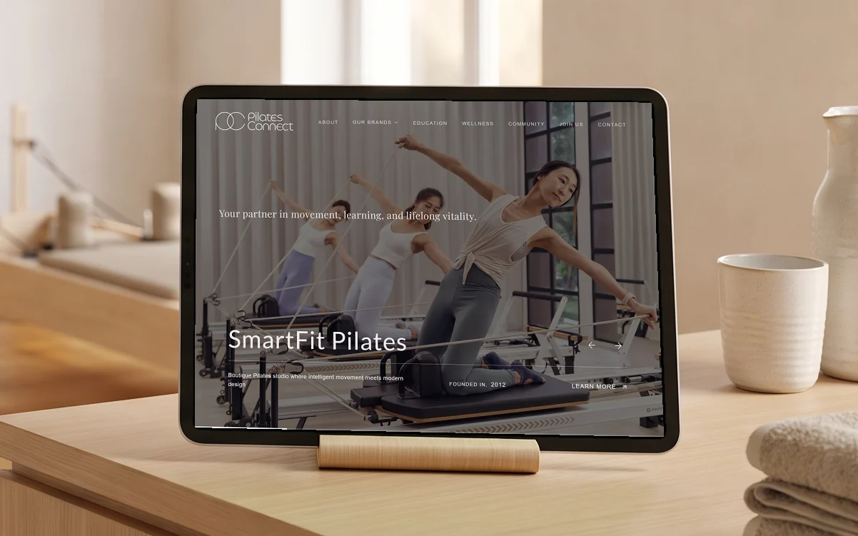 Pilates Connect homepage concept, "Your partner in movement, learning, and lifelong vitality." over the SmartFit Pilates slide of a reformer class, on a tablet on a pale oak stand on the light-wood reception counter of a pilates studio, reformers soft in the morning light behind