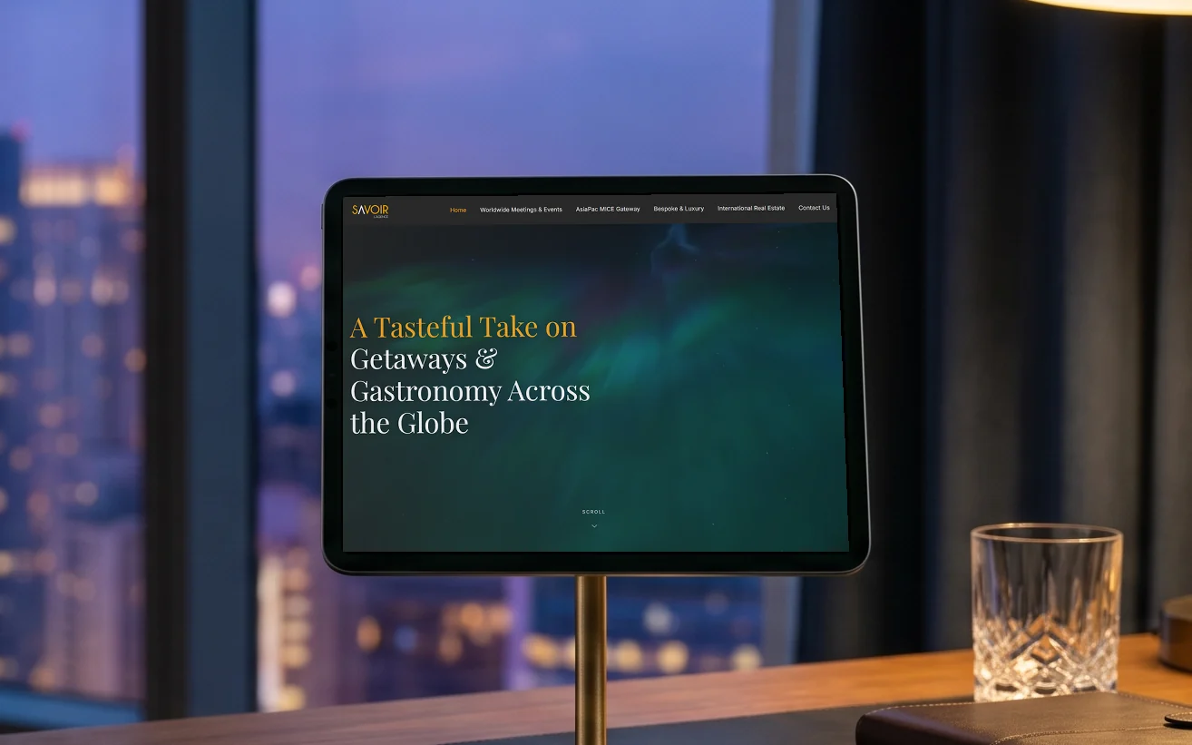 SAVOIR L'Agence homepage, "A Tasteful Take on Getaways & Gastronomy Across the Globe" over the northern lights, on a tablet on a brass stand in a hotel suite at dusk