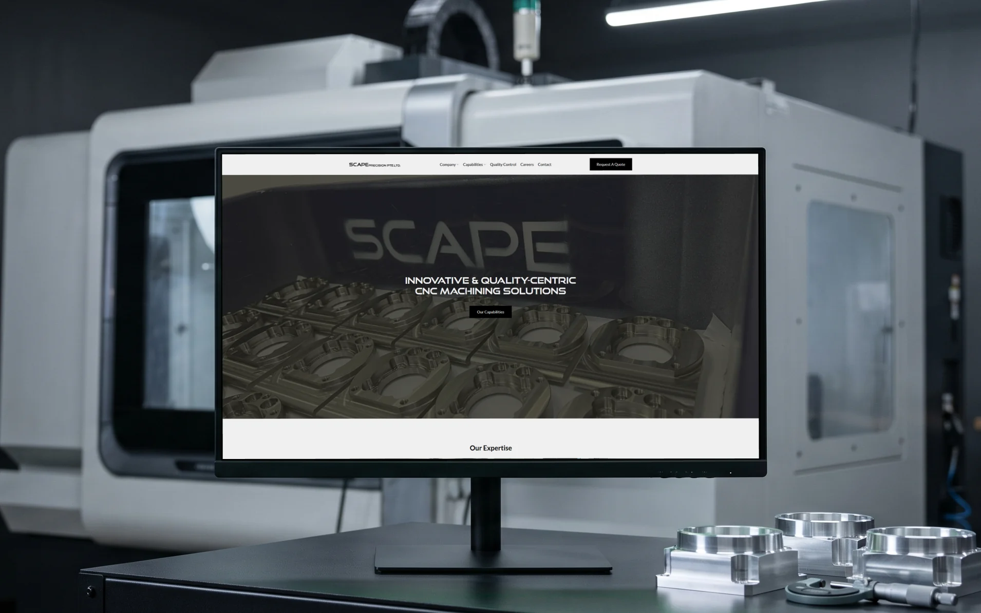 Scape Precision homepage, "Innovative & Quality-Centric CNC Machining Solutions" over a photograph of machined housings, on a 27-inch monitor on an industrial desk beside a CNC machine and machined aluminium parts