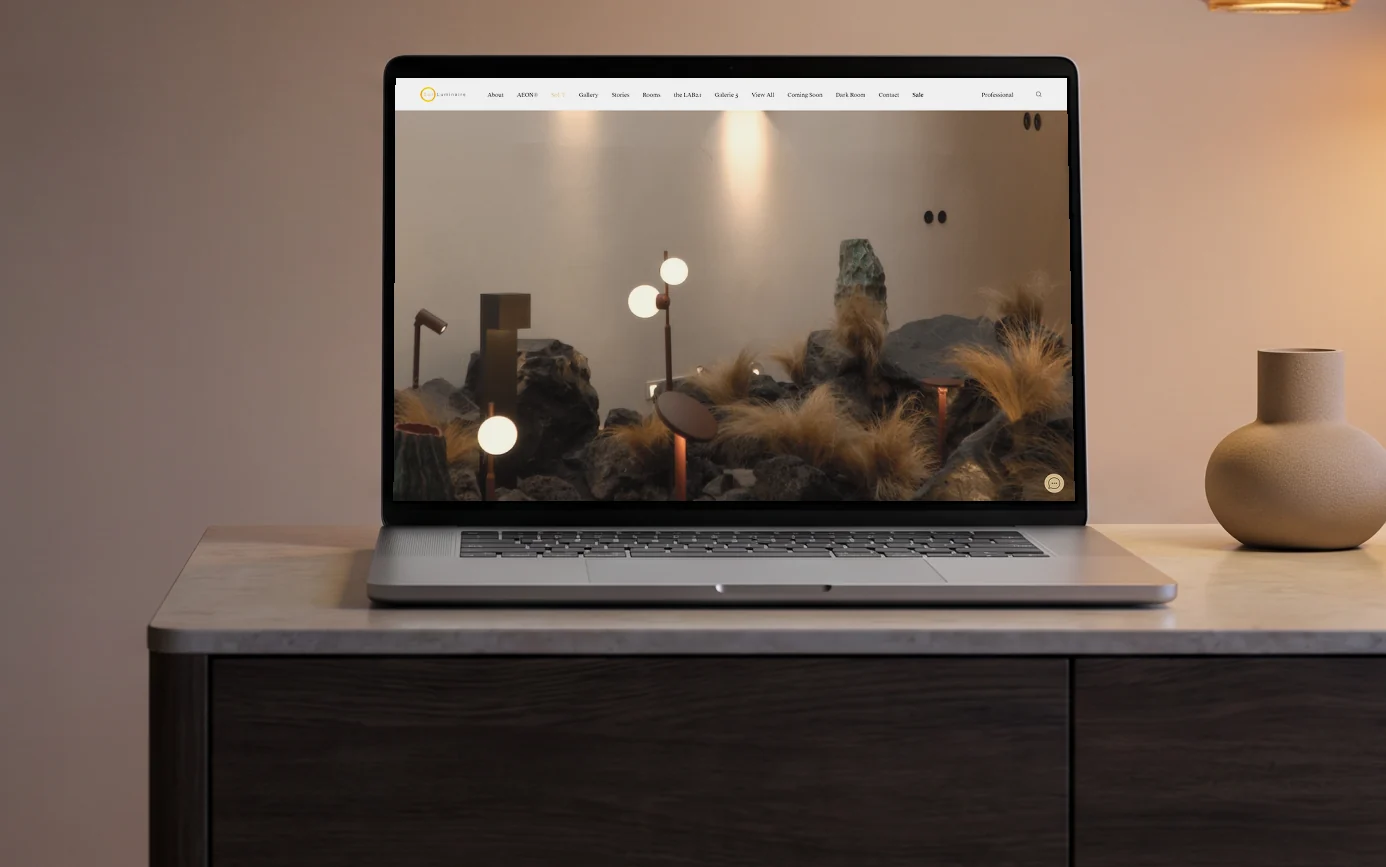 Sol Luminaire homepage, its showroom film of glowing globe lights over a volcanic rock and feather-grass garden, on a laptop on a smoked-oak sideboard under two glowing designer pendant lamps at dusk
