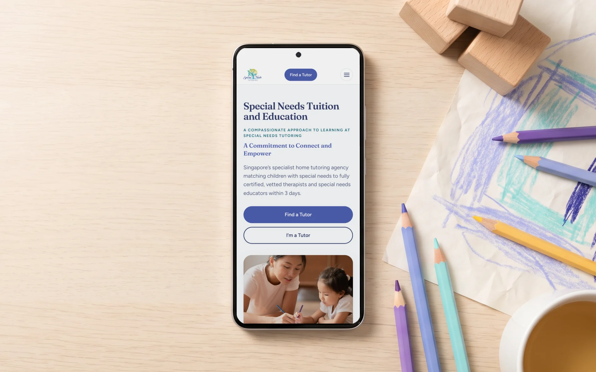 Special Needs Tutoring mobile site, "Special Needs Tuition and Education" with its Find a Tutor button, on a phone lying on an oak table beside a child's drawing and coloured pencils