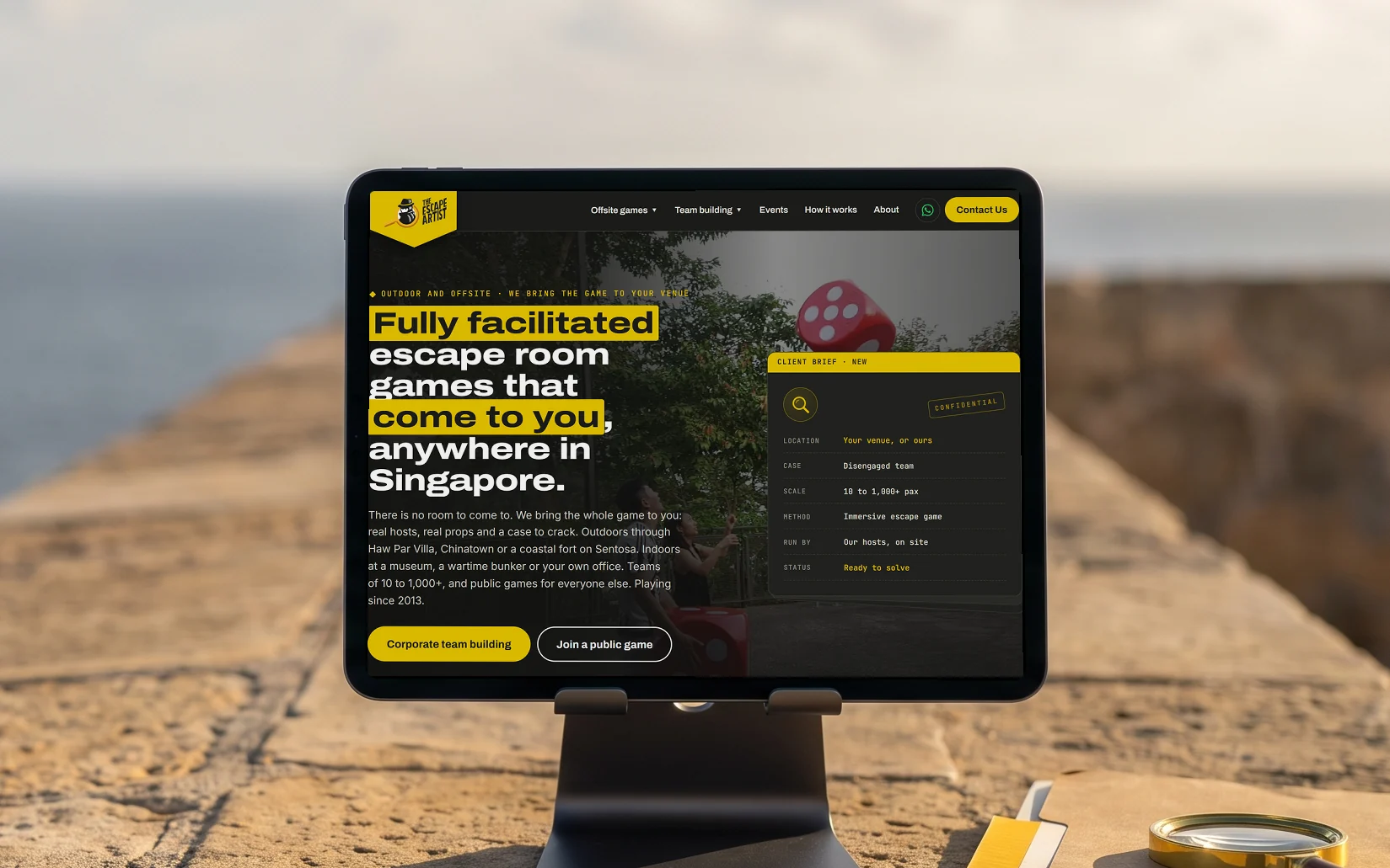 The Escape Artist homepage, "Fully facilitated escape room games that come to you, anywhere in Singapore.", on a tablet on a stand on the stone parapet of an old coastal fort in late-afternoon sun, the sea behind and a case file with a brass magnifying glass beside it