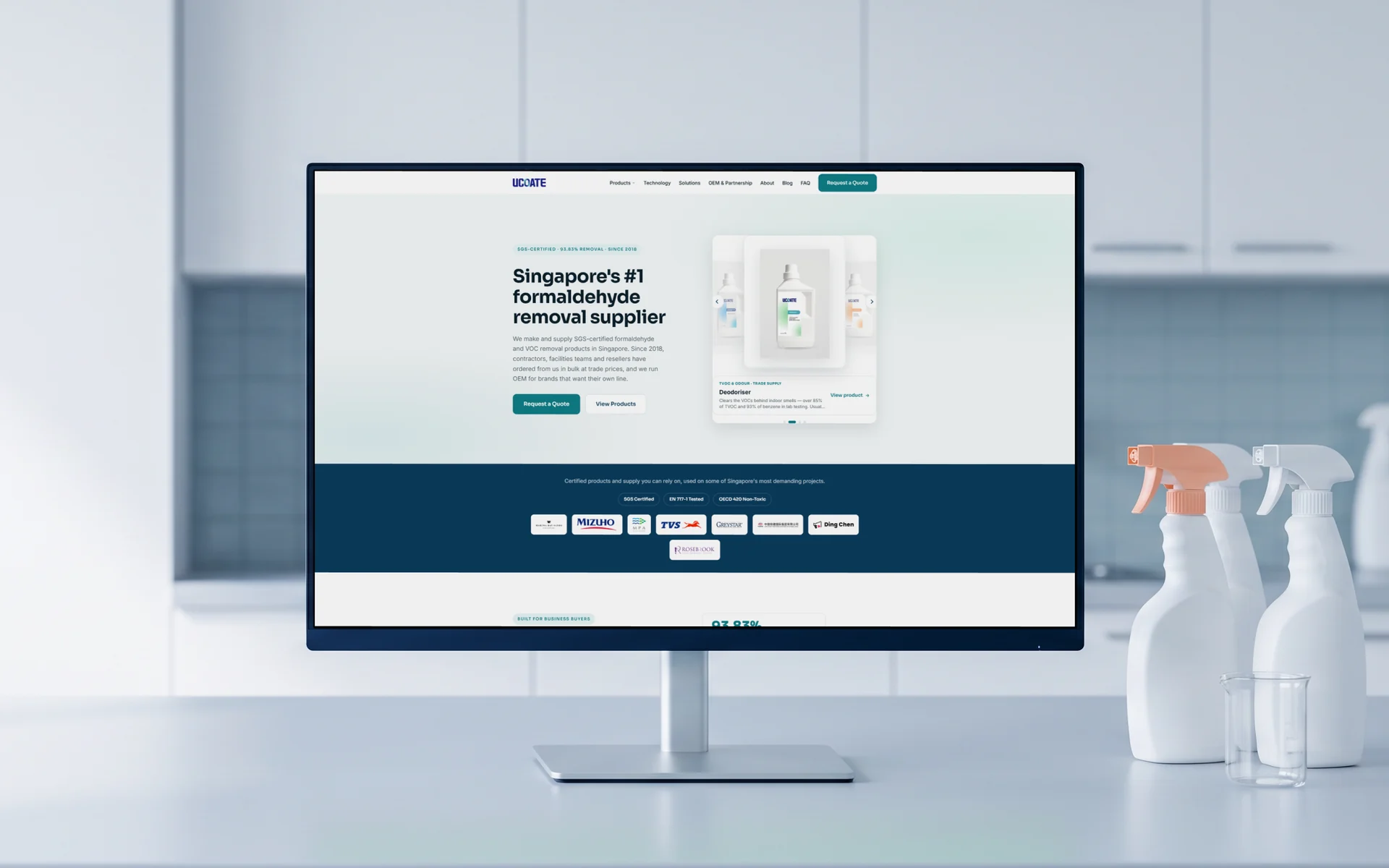 UCOATE homepage, "Singapore's #1 formaldehyde removal supplier" above its SGS Certified, EN 717-1 Tested and OECD 420 Non-Toxic band, on a monitor on a pale lab counter in a bright testing room, unlabelled spray bottles and a glass beaker beside it