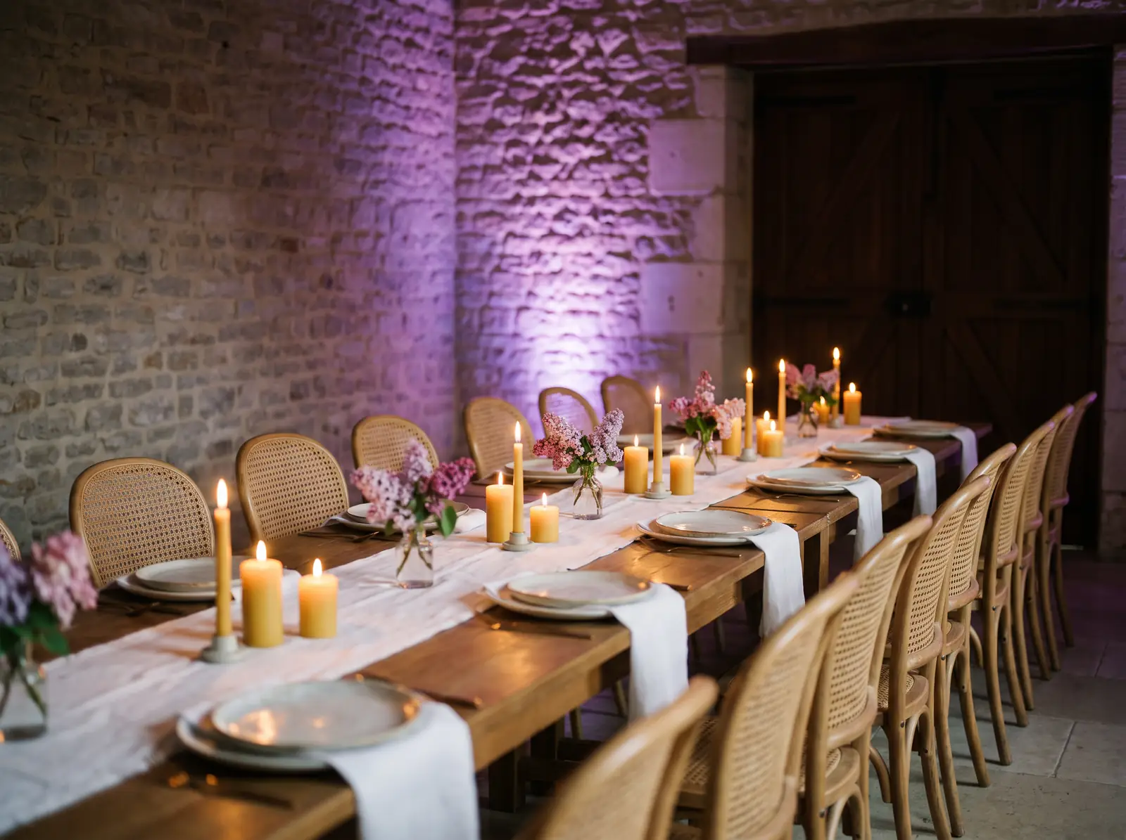 A long candlelit dinner table with ceramic plates and lilac flowers, purple light on the stone wall behind