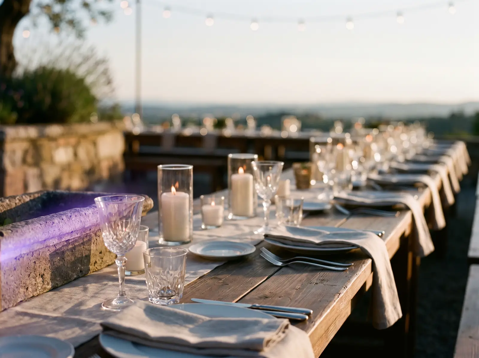 A long candlelit table set for dinner on a stone terrace at sunset